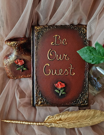 A decorative vintage-style book with ornate gold lettering that reads 'Be Our Guest' on a dark reddish-brown cover, styled with draped blush fabric, a small vase, a glass bottle, and a gold feather quill.