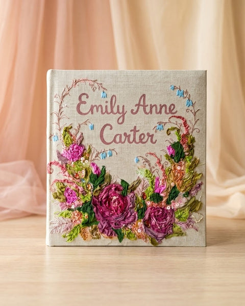 A square fabric keepsake box with custom embroidery, featuring the name 'Emily Anne Carter' in pink script surrounded by a floral wreath and pink roses, set on a light wooden surface with soft beige draped fabric in the background.