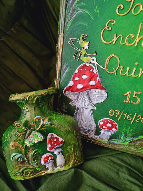 A close-up of a green decorative plaque or keepsake board featuring painted fairy and mushroom illustrations, placed beside a matching green ornament or box piece on draped dark green fabric.