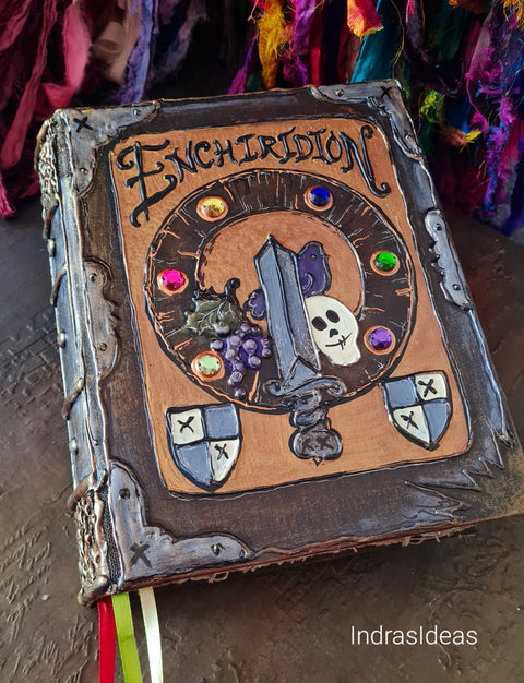 A decorative gothic-style book titled 'Enchiridion' with a brown leather-like cover, ornate dark metal corners, a central illustration of a sword, skull, grapes, and shields, and colorful gem accents. The book is photographed on a dark surface with colorful fabric in the background.