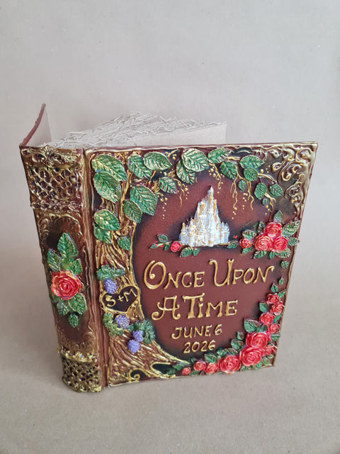 An ornate decorative book-shaped keepsake box with embossed floral details, gold lettering reading 'Once Upon a Time June 6 2026,' and a small castle motif on the front, photographed against a plain light gray background.