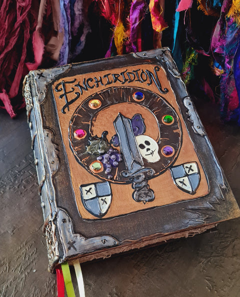 A handcrafted fantasy 'Enchiridion' journal with a medieval-themed cover featuring a sword, skull, gems, and shields, resting on a textured surface with colorful ribbons and hanging textiles in the background.