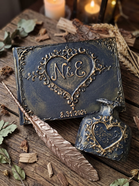 A decorative keepsake book and matching ink bottle with embossed hearts and the initials 'N&E' on a rustic wooden surface, styled with dried leaves, a feather quill, and blurred candles in the background.