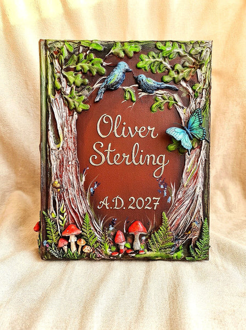 A personalized 3D raised book cover plaque featuring a woodland theme with two blue birds, a blue butterfly, red mushrooms, and green leaves framing the name 'Oliver Sterling' and the date 'A.D.