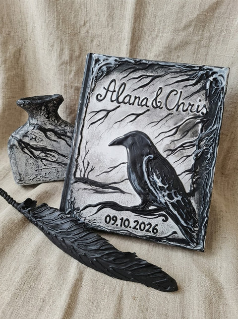 A gothic-themed keepsake box set displayed on a beige linen fabric background. The set includes a square metal box engraved with a raven, tree branches, and the personalized names 'Alana & Chris' along with the date '09.