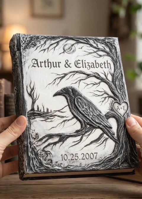 A personalized hardcover journal held by two hands, featuring a detailed black and white illustration of a raven perched on a tree branch with the names 'Arthur & Elizabeth' and the date '10.25.