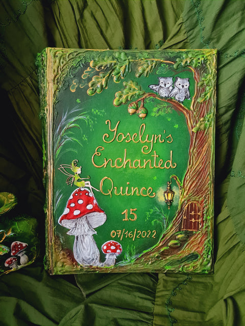 A personalized hardcover journal titled 'Yosclyn's Enchanted Quince' featuring whimsical hand-painted forest illustrations including a fairy, red-and-white mushrooms, a tree with two cats, and a small house, displayed on a textured green fabric background.