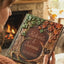 A person holds a personalized embossed Autumn Wedding guest book in front of a cozy fireplace. The album features a detailed 3D design of tree roots and branches with green ivy and orange autumn leaves, and the names 'Robert & Tammi' and the date '26/10/2027' are engraved in the center