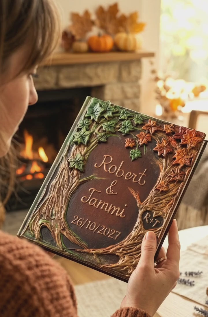 A person holds a personalized embossed Autumn Wedding guest book in front of a cozy fireplace. The album features a detailed 3D design of tree roots and branches with green ivy and orange autumn leaves, and the names 'Robert & Tammi' and the date '26/10/2027' are engraved in the center
