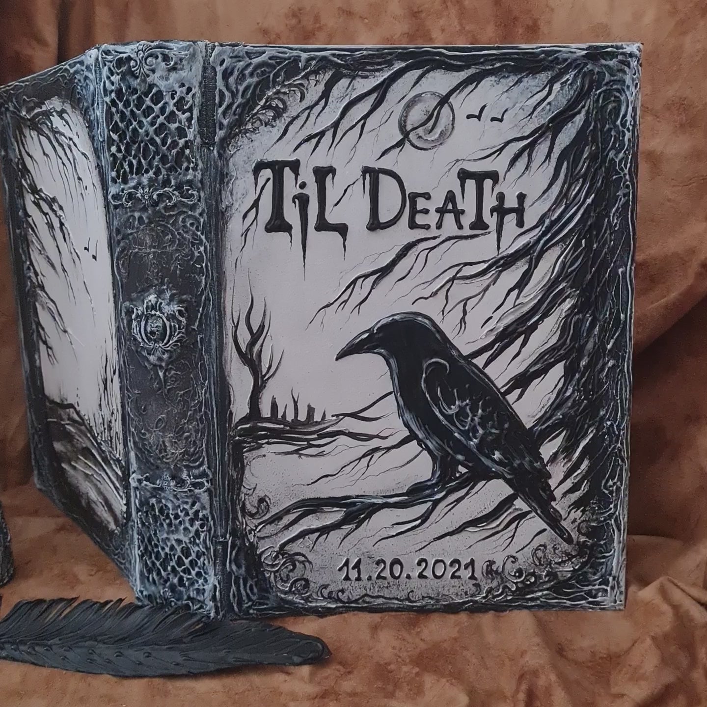 A gothic-style hardcover  handmade journal or notebook with a 3D raven illustration and the text 'Til Death' on the cover, photographed on a brown textured fabric background.