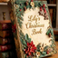 Personalized handmade Christmas memory book with 3D sculpted relief cover featuring gingerbread man and star ornaments, fir boughs, and text "Lily's Christmas Book" on an antique cream cover, resting on a wooden desk with candles.