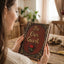A woman in a cream knit sweater holds a decorative brown leather-bound book or journal with gold lettering that reads 'Be Our Guest' and a small red rose on the cover, set in a warm, softly lit room.