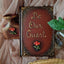 A decorative vintage-style book with ornate gold lettering that reads 'Be Our Guest' on a dark reddish-brown cover, styled with draped blush fabric, a small vase, a glass bottle, and a gold feather quill.