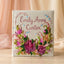 A square fabric keepsake box with custom embroidery, featuring the name 'Emily Anne Carter' in pink script surrounded by a floral wreath and pink roses, set on a light wooden surface with soft beige draped fabric in the background.