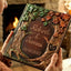 A person holds a personalized. Embossed Fall wedding guest book in front of a cozy fireplace. The album features a detailed 3D design of tree roots and branches with green ivy and orange autumn leaves, and the names 'Robert & Tammi' and the date '26/10/2027' are engraved in the center