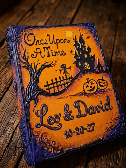 A personalized Halloween-themed keepsake journal with a textured orange and purple cover, featuring embossed illustrations of a haunted castle, a spooky tree, pumpkins, and the text 'Once Upon A Time Leo & David 10-20-27', displayed on a rustic wooden surface. IndrasIdeas