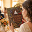 A woman in a champagne lace gown holds a custom wedding guest book featuring a dark brown cover decorated with purple grapes, red roses, and gold lettering that reads 'Once Upon A Time September 25 2026'. The background shows a softly blurred, warmly lit wedding reception venue with tables and guests.