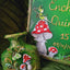 A close-up of a green decorative plaque or keepsake board featuring painted fairy and mushroom illustrations, placed beside a matching green ornament or box piece on draped dark green fabric.