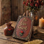 A rustic decorative sign reading 'Be Our Guest' displayed on a small easel stand on a wooden table, styled with dried red roses, a small vase, a candle, and a gold feather quill against a wood plank background.