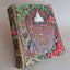 A handcrafted decorative book or journal with an ornate embossed cover reading 'Once Upon a Time June 6 2026,' featuring green leaves, red roses, purple grapes, gold lettering, and a small white castle motif on a neutral background.