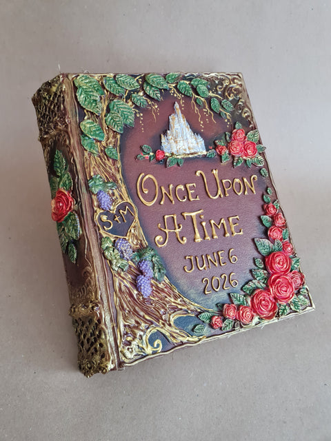 A handcrafted decorative book or journal with an ornate embossed cover reading 'Once Upon a Time June 6 2026,' featuring green leaves, red roses, purple grapes, gold lettering, and a small white castle motif on a neutral background.