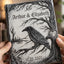 A personalized gothic-style hardcover journal held by two hands. The cover features a detailed black ink illustration of a raven perched on a bare tree branch under a full moon, with the names 'Arthur & Elizabeth' and the date '10.