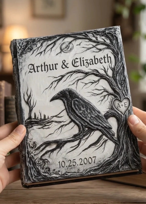 A personalized gothic-style hardcover journal held by two hands. The cover features a detailed black ink illustration of a raven perched on a bare tree branch under a full moon, with the names 'Arthur & Elizabeth' and the date '10.