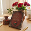 A decorative leather-style sign or notebook with the words 'Be Our Guest' in gold lettering, displayed on a wooden table with red roses in a vase, a small vase, and a gold pen nearby.
