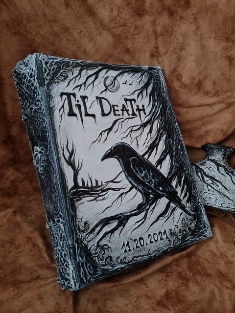 A gothic-style hardcover journal titled 'Til Death' featuring a black raven perched on tree branches over a dark landscape, resting on a brown textured fabric background.