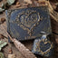 A decorative keepsake book and matching ink bottle with embossed hearts and the initials 'N&E' on a rustic wooden surface, styled with dried leaves, a feather quill, and blurred candles in the background.
