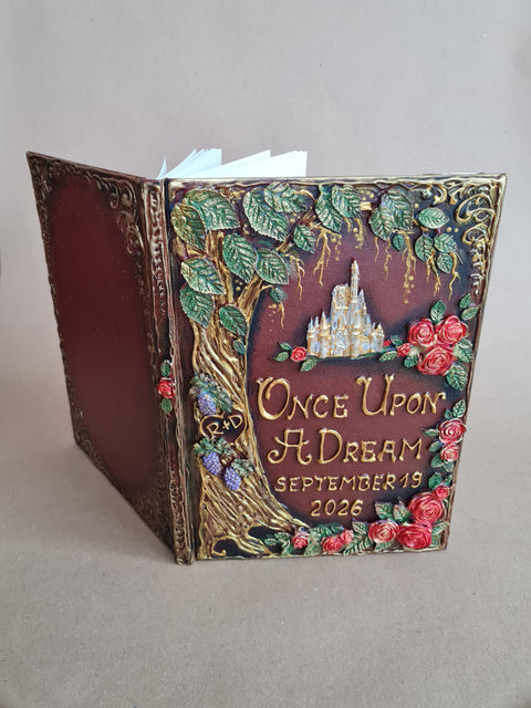 A decorative hardcover journal standing open on a neutral gray background. The cover features embossed floral and tree details with red roses, green leaves, and gold lettering that reads 'Once Upon A Dream September 19 2026' with a small castle illustration above.