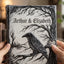 A personalized hardcover journal held by two hands, featuring a black and white gothic illustration of a raven perched on a tree branch with the names 'Arthur & Elizabeth' and the date '10.25.
