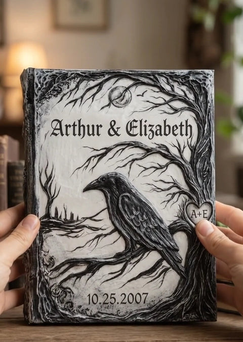 A personalized hardcover journal held by two hands, featuring a black and white gothic illustration of a raven perched on a tree branch with the names 'Arthur & Elizabeth' and the date '10.25.