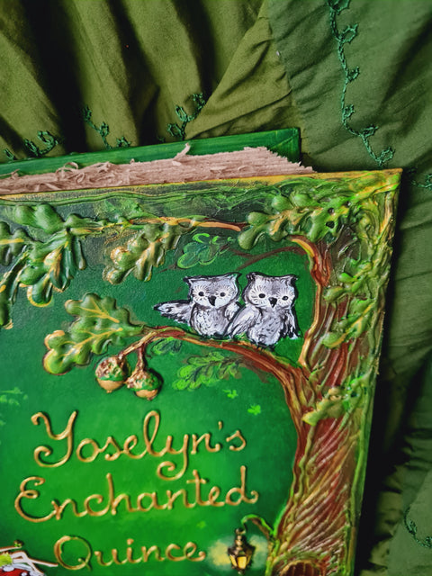 A close-up of a decorative book cover titled "Yoselyn's Enchanted Quince" featuring two gray owls perched on a tree branch with green leaves and brown trunk, set against a dark green embroidered fabric background.
