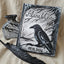A gothic-themed keepsake box set displayed on a beige linen fabric background. The set includes a square metal box engraved with a raven, tree branches, and the personalized names 'Alana & Chris' along with the date '09.