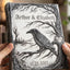 A personalized hardcover journal held by two hands, featuring a detailed black and white illustration of a raven perched on a tree branch with the names 'Arthur & Elizabeth' and the date '10.25.