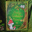 A personalized hardcover journal titled 'Yosclyn's Enchanted Quince' featuring whimsical hand-painted forest illustrations including a fairy, red-and-white mushrooms, a tree with two cats, and a small house, displayed on a textured green fabric background.